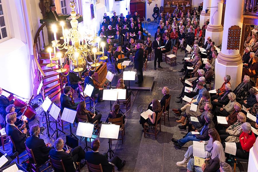 Overzicht van de kerk tijdens de uitvoering: het publiek rechts met programmaboekjes op schoot, het orkest links, en een brandende kroonluchter boven de zaal.
